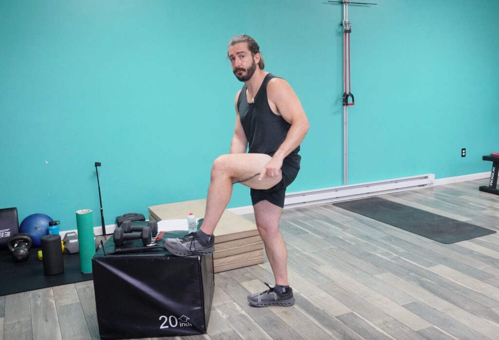 Dr. John standing with his left foot elevated on a box. He is holding a bow muscle scraper in his hands and is applying pressure to his hamstrings with it as a form of pulled hamstring treatment
